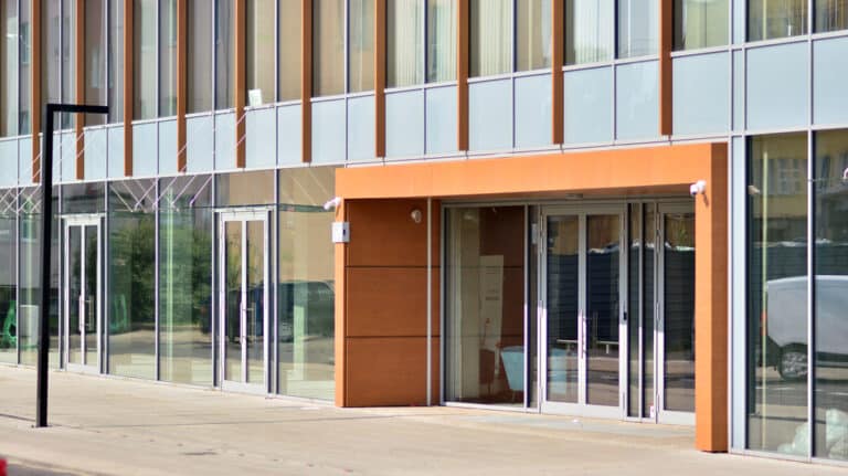 Modern office building exterior with glass curtain walls, aluminium cladding panels, and a central orange-panelled entrance.